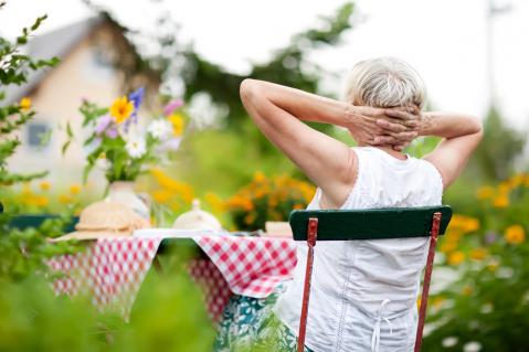 Ältere Frau sitzt auf einem Stuhl in ihrem Garten und schaut auf ein Haus